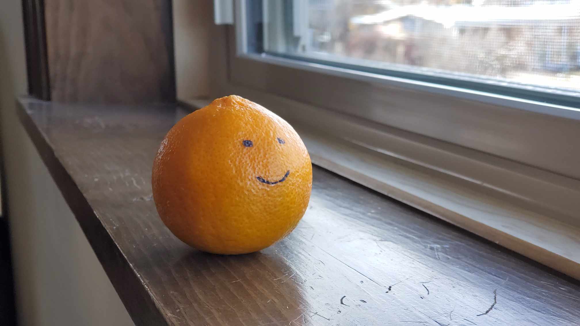 A mandarin orange with a face drawn on it photographed on a window sill.